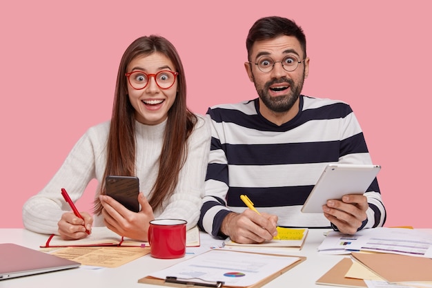 Teamarbeiter sitzen an einem Schreibtisch, machen sich Notizen im Notizblock, bereiten sich auf das Seminar vor, halten ein modernes Smartphone und Touchpad in der Hand, haben einen glücklichen Blick, studieren Dokumente mit Diagrammen und sind in den Arbeitsprozess involviert