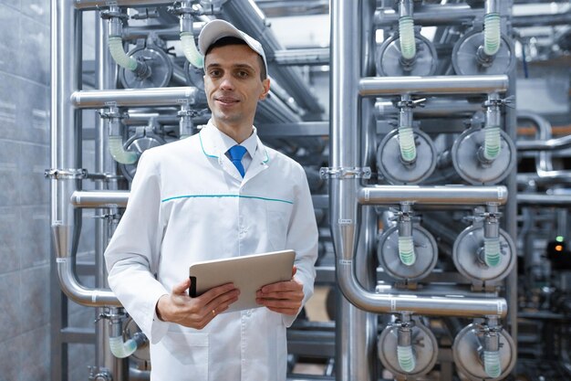 Technologe in einem weißen Mantel macht die notwendigen Eingaben in das Tablet ist in der Fabrik Ein Mann mit einem digitalen Tablet in Uniform in der Produktionshalle der Produktionsabteilung im Hintergrund