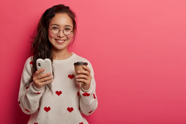 Technologie-, Kommunikations- und Lifestyle-Konzept. Hübsches junges Mädchen in runden Gläsern benutzt Smartphone zum Lesen von coolen Blogs und Nachrichten, trinkt Kaffee zum Mitnehmen, lädt App herunter, steht drinnen.