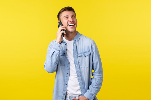 Technologie, Lifestyle-Konzept. Fröhlicher, gutaussehender lächelnder Mann, der ein glückliches Gespräch am Telefon führt, einen Freund anruft, begeistert aufschaut und Smartphone in der Nähe des Ohrs hält, gelber Hintergrund.