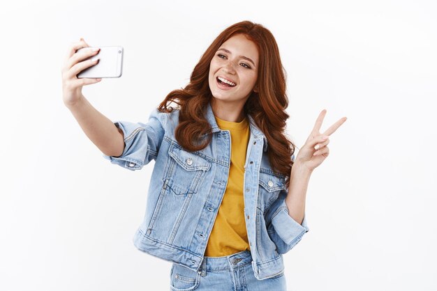 Technologie-Lifestyle- und Frauenkonzept Fröhliches, süßes, stilvolles Mädchen mit rotem Kopf verlängert die Hand mit dem Smartphone, das ein Selfie macht, das sorglos lächelt und das Siegeszeichen des Friedens auf weißem Hintergrund zeigt