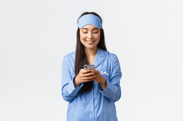 Technologie, Menschen und Freizeitkonzept. Lächelnde süße asiatische Bloggerin im Pyjama und Schlafmaske, Post in sozialen Medien vor dem Schlafen schreiben, Nachrichten mit dem Handy