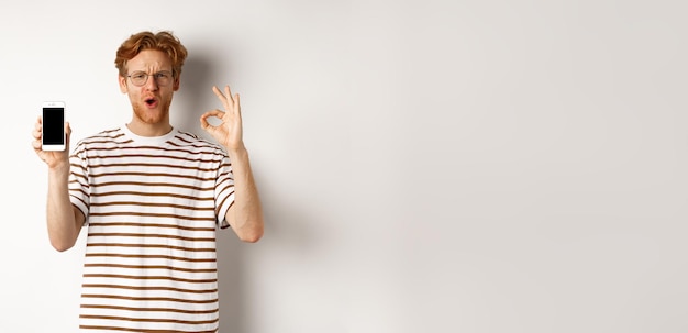 Kostenloses Foto technologie- und e-commerce-konzept junger mann mit roten haaren, der ein okay-schild und einen leeren smartphone-scre zeigt