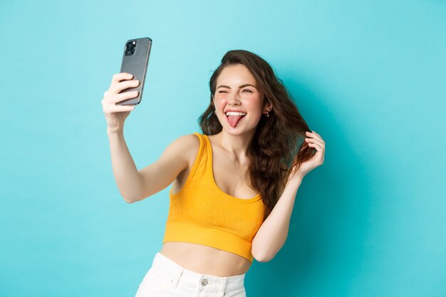 Technologie- und Lifestyle-Konzept. Glückliche junge Frau, die dumme Gesichter macht, während sie Selfie auf Smartphone-App mit Filtern macht und vor blauem Hintergrund steht.
