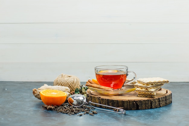 Tee in einer Tasse mit Kräutern, Orange, Gewürzen, Waffel, Faden, Holzbrett, Siebseitenansicht auf weißem und Stuckhintergrund