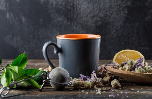 Tee in einer Tasse mit Limetten, Sieb, Kräutern, Zitrone, Limetten-Seitenansicht auf dunkler und hölzerner Oberfläche