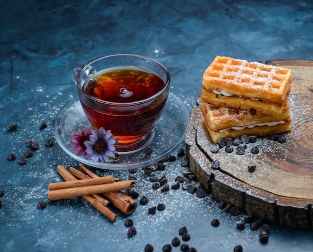 Tee in einer Tasse mit Zimtstangen, Waffel, Schokostückchen, Blumen, Holzbrett-Hochwinkelansicht auf einer blauen Oberfläche