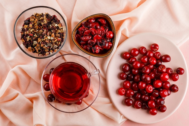 Tee mit Kirschen, Marmelade, getrockneten Kräutern in einem Glasbecher auf Pink und Textil.