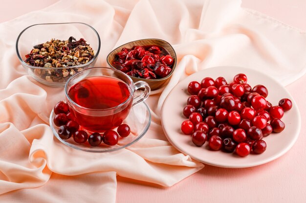 Tee mit Kirschen, Marmelade, getrockneten Kräutern in einem Glasbecher auf rosa und textil hoch.