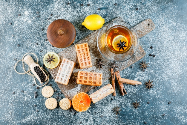 Tee mit Waffel, Keks, Gewürzen, Choco-Chips, Orange in einer Tasse auf grungy und Schneidebrettoberfläche, flach legen.