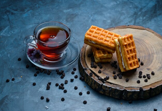 Tee mit Waffel, Schokoladenstückchen, Blumen in einer Tasse auf blauer und hölzerner Brettoberfläche, hohe Winkelansicht.