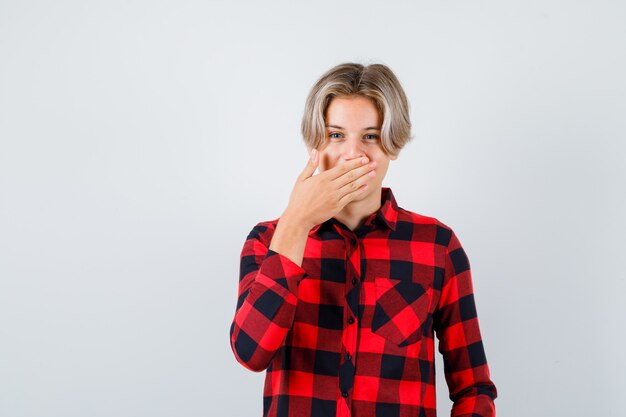 Teen blonder Mann in Freizeithemd, der den Mund mit der Hand bedeckt und fröhlich aussieht, Vorderansicht.