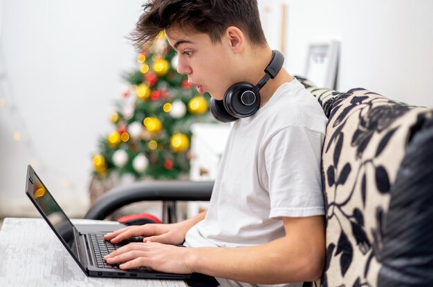 Teen Boy benutzt Laptop mit Kopfhörern zu Hause. Weihnachtsbaum an der Wand. Erstauntes Gesicht