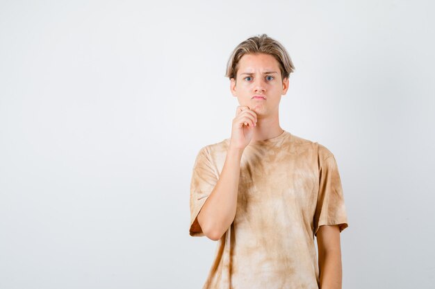 Teen Boy hält Hand am Kinn im T-Shirt und sieht nachdenklich aus. Vorderansicht.