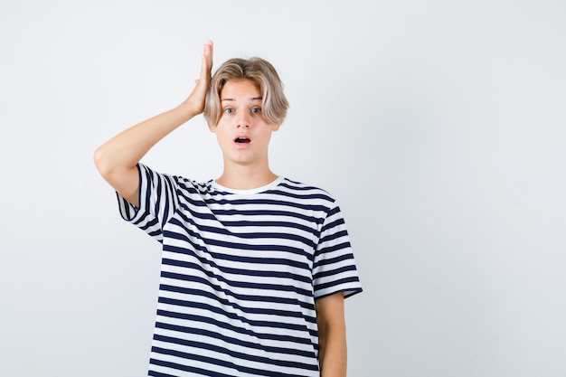 Teen Boy im T-Shirt, das die Hand auf dem Kopf hält, während er den Mund öffnet und schockiert aussieht, Vorderansicht.