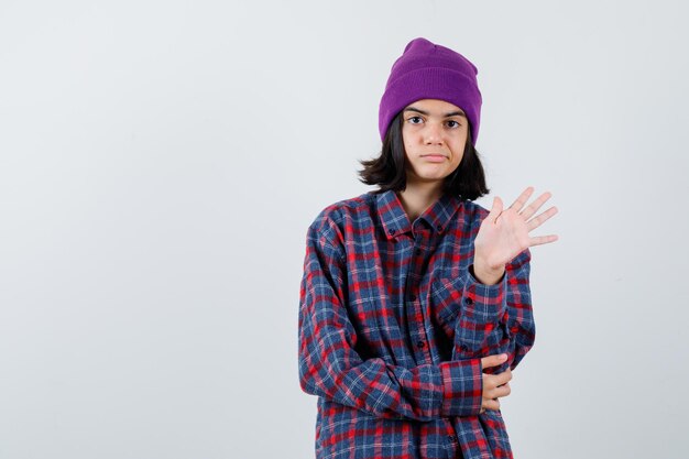Teen Frau winkt mit der Hand in kariertem Hemd lila Beanie zu grüßen, die liebenswürdig aussieht