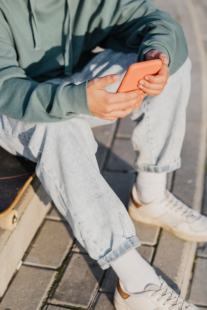 Teenager außerhalb, der Smartphone beim Sitzen auf Skateboard hält