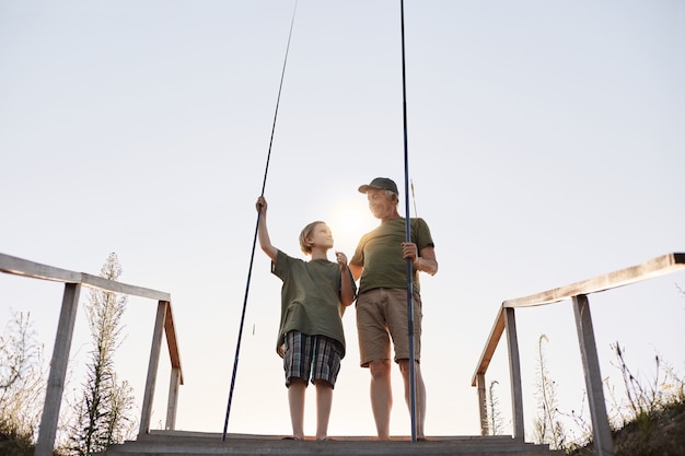 Teenager, der lernt, mit angelrute zu fischen, opa lehrt seinen enkel, fische zu fangen, porträt in voller länge auf hölzernem ponton mit treppen, schöner sonnenuntergang.