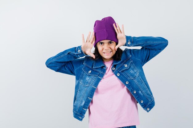 Teenager-Frau, die erhobene Hände in der Nähe des Kopfes in der T-Shirt-Jackenmütze hält, die lustig aussieht