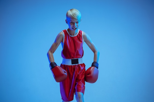 Teenager im Sportbekleidungsboxen lokalisiert auf blauem Studiohintergrund im Neonlicht. Anfänger männlicher kaukasischer Boxer, der hart trainiert und trainiert, tritt. Sport, gesunder Lebensstil, Bewegungskonzept.