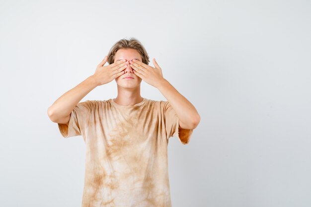 Teenager-Junge, der die Augen mit den Händen im T-Shirt bedeckt und verängstigt aussieht, Vorderansicht.