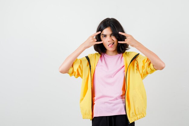 Teenager-Mädchen, das Rock n Roll-Geste zeigt, die Zunge in T-Shirt, Jacke herausstreckt und fröhlich aussieht, Vorderansicht.