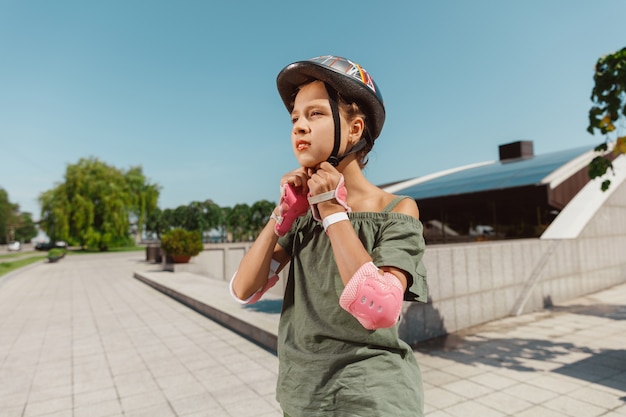 Teenager-Mädchen in einem Helm lernt, auf Rollschuhen zu fahren, die eine Balance halten oder Inlineskaten und sich an einem sonnigen Sommertag auf der Straße der Stadt drehen