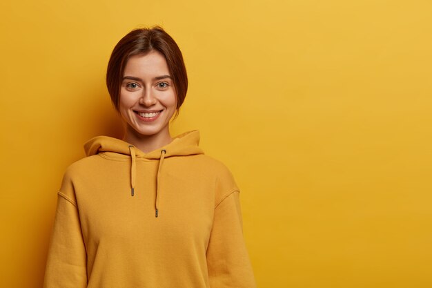 Teenager-Mädchen mit angenehmem Lächeln, hat dunkles gekämmtes Haar, trägt Sweatshirt, hat gesunde Haut, steht gegen gelben Raum