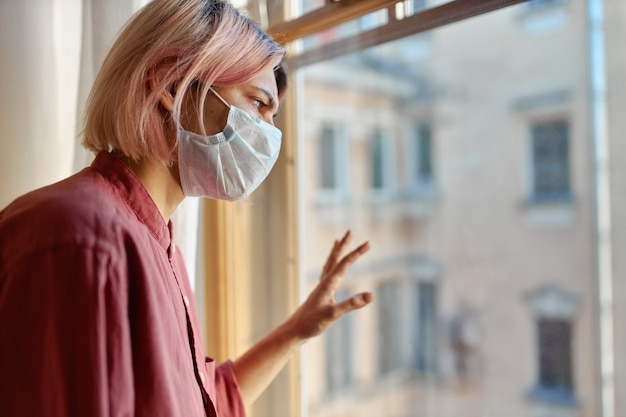 Kostenloses Foto teenager-mädchen mit rosa haaren, das vor geschlossenem fenster mit hand auf glas steht und nach draußen schaut, während zu hause während der quarantäne bleibt. coronavirus-pandemie und soziales distanzierungskonzept