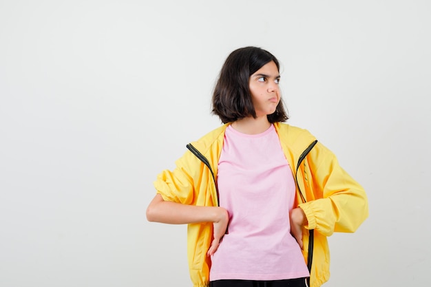 Teenie-mädchen im gelben trainingsanzug, t-shirt, das die hände an der taille hält, zur seite schaut und unzufrieden aussieht, vorderansicht.