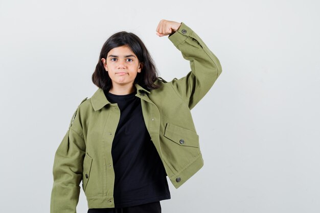 Teenie-Mädchen im T-Shirt, grüne Jacke, die Armmuskeln zeigt und selbstbewusst aussieht, Vorderansicht.