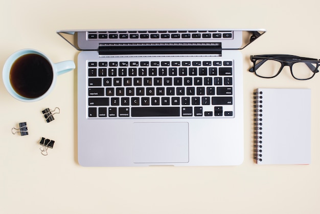 Teetasse; Büroklammern; Brille; Laptop und gewundenes Notizbuch auf beige Hintergrund