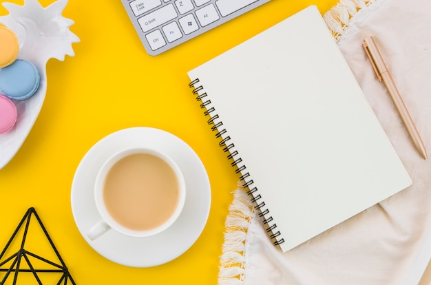 Teetasse; Makronen; Spiralblock; Stift auf Tischdecke vor gelbem Hintergrund