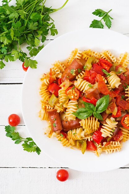 Teigwaren mit Tomatensauce mit Wurst, Tomaten, grüner Basilikum verziert in der weißen Platte auf einem Holztisch.