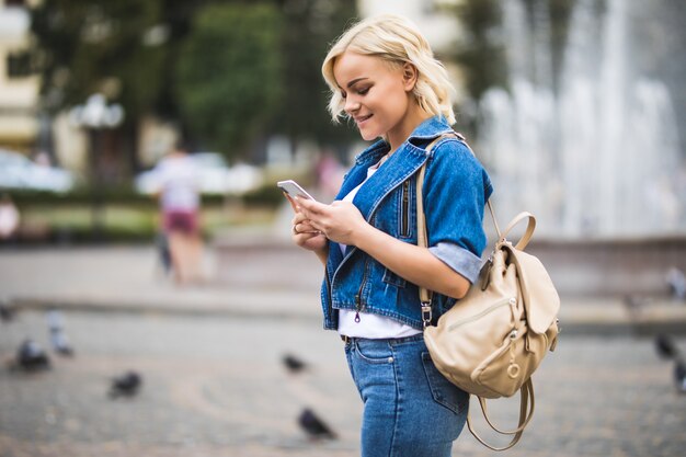 Telefon der jungen blonden Mädchenfrau in ihren Händen auf Straßenwegquadrat-Fontain, gekleidet in Blue Jeans Suite mit Tasche auf ihrer Schulter in sonnigem Tag