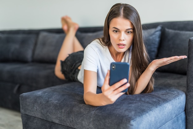 Telefonbenutzer der jungen Frau, die Videoanruf hat. Junge Frau im lässigen Liegen auf Couch, mit Smartphone und Lächeln am Bildschirm.