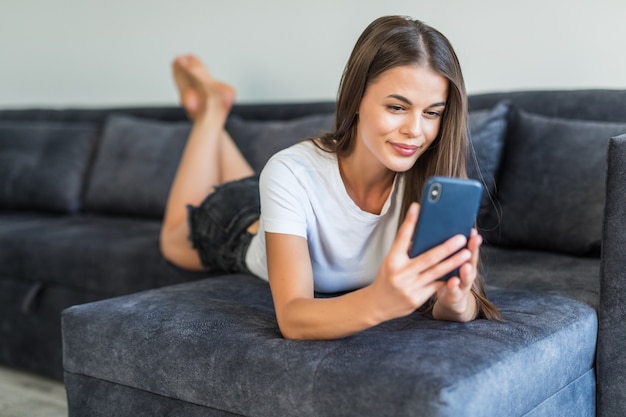 Telefonbenutzer der jungen Frau, die Videoanruf hat. Junge Frau im lässigen Liegen auf Couch, mit Smartphone und Lächeln am Bildschirm.