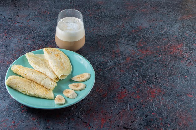 Teller mit Crpes und Bananenscheiben mit Glas Milchkaffee auf dunkler Oberfläche.