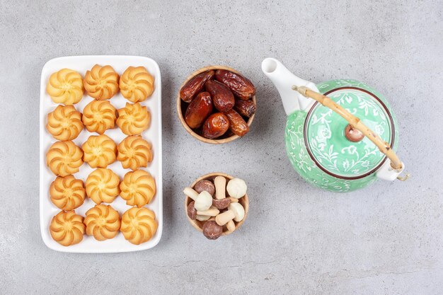 Teller mit Keksen neben verzierter Teekanne und Schalen mit Datteln und Pilzschokoladen auf Marmorhintergrund. Hochwertiges Foto