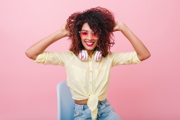 Terrific afrikanisches Mädchen in der rosa Sonnenbrille, die im blauen Stuhl sitzt. Lachendes schwarzes weibliches Modell im Baumwollhemd, das mit lockigem braunem Haar spielt.