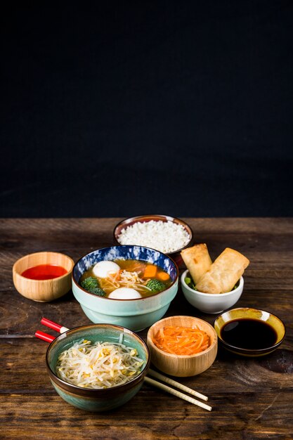 Thailändische Fischballsuppe; Frühlingsrollen; Bohnen sprießen und Sauce mit Stäbchen auf hölzernen Schreibtisch vor schwarzem Hintergrund