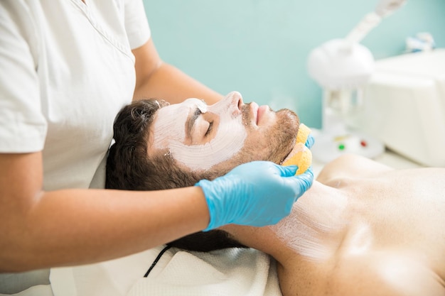 Therapeutin wäscht einem männlichen Kunden das Gesicht, während sie in einem Spa eine Gesichtsbehandlung gibt