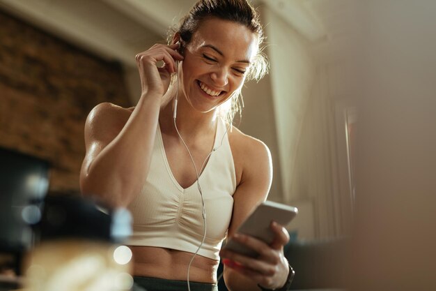 Tiefwinkelansicht einer glücklichen sportlichen Frau, die ihre Kopfhörer anpasst, während sie zu Hause Musik auf dem Smartphone hört