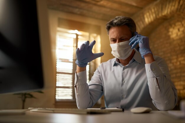 Tiefwinkelansicht eines Geschäftsmannes mit Gesichtsmaske und Handschuhen, der während der Coronavirus-Epidemie über das Handy kommuniziert, während er im Büro arbeitet