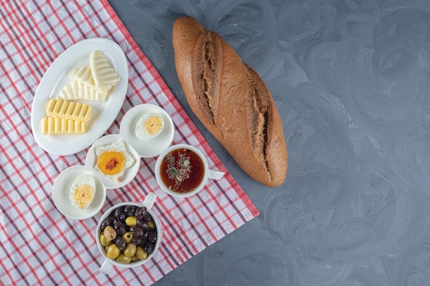 Tischdecke unter einem Frühstücksbündel Brotlaib und Platten mit Käse, Butter und Ei, mit Tee und Oliven auf Marmorhintergrund.