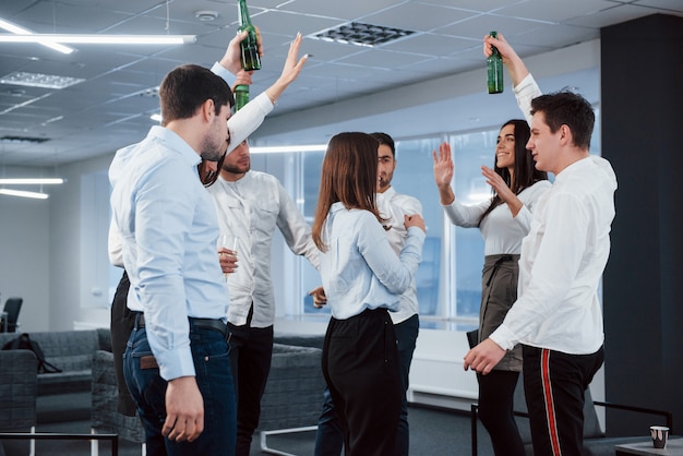 Toast machen. Foto des jungen Teams in der klassischen Kleidung, die Erfolg feiert, während Getränke im modernen gut beleuchteten Büro halten