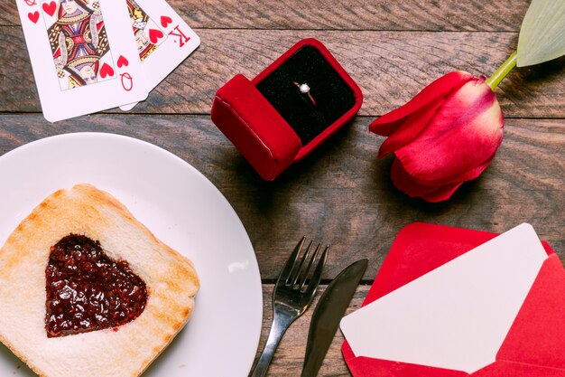 Toast mit Marmelade auf Teller in der Nähe von Spielkarten, Blume, Umschlag und Ring in Geschenkbox