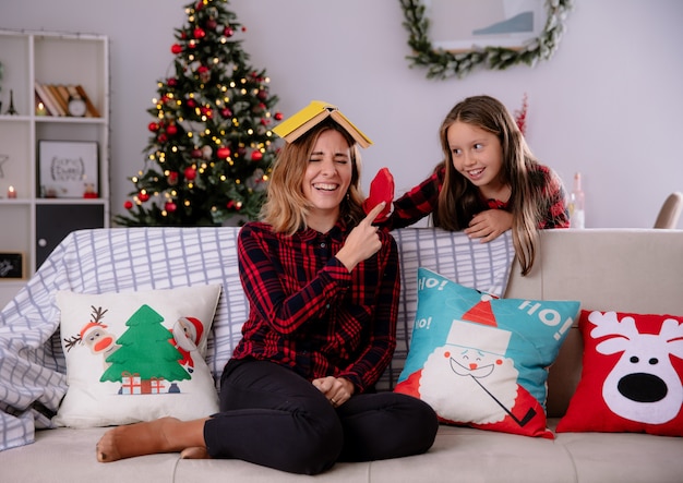 Tochter spielt mit Strümpfen, die hinter ihrer Mutter stehen und Buch auf Kopf halten, auf Couch sitzen und Weihnachtszeit zu Hause genießen
