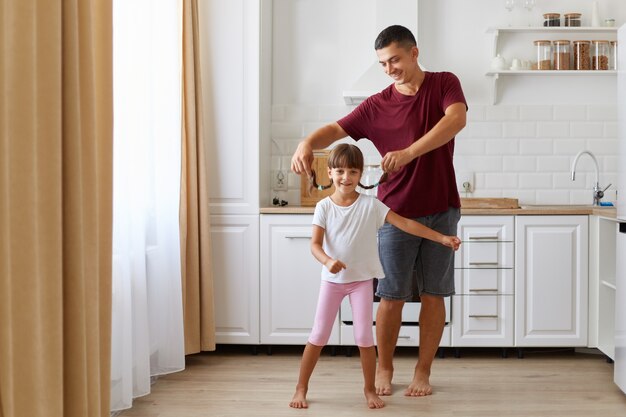 Tochter und Vater, die Spaß haben und in der Küche tanzen, Leute, die Freizeitkleidung tragen, Mann, der kleine Mädchenzöpfe aufzieht, glückliche Familie, die Zeit zu Hause zusammen verbringt.