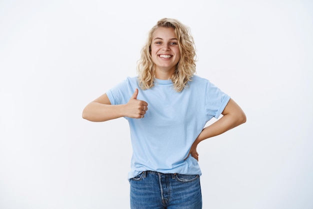 Tolle, gute Arbeit. Porträt einer grinsenden, süßen und freundlichen, charismatischen, attraktiven Blondine mit blauen Augen, die erfreut lächelt, als Daumen hochzustimmend und wie, positive Meinung zur weißen Wand gebend.